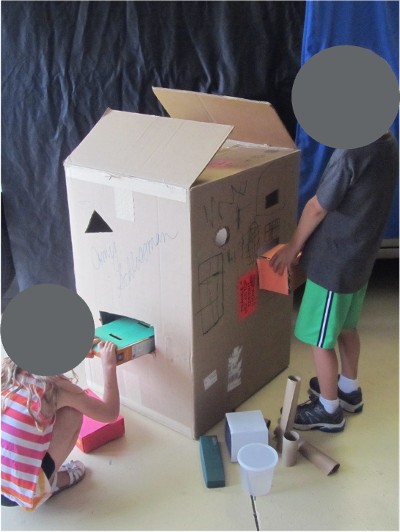 Two children place items in a large recycled box through the cut holes