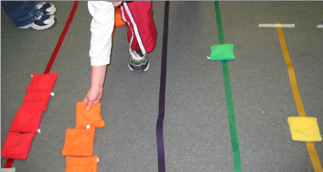 A child lays square colored bean bags to match the colored ribbons on the floor