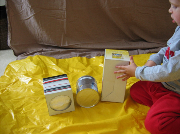 Child seated on yellow tarp playing with two recycled boxes and one can. 